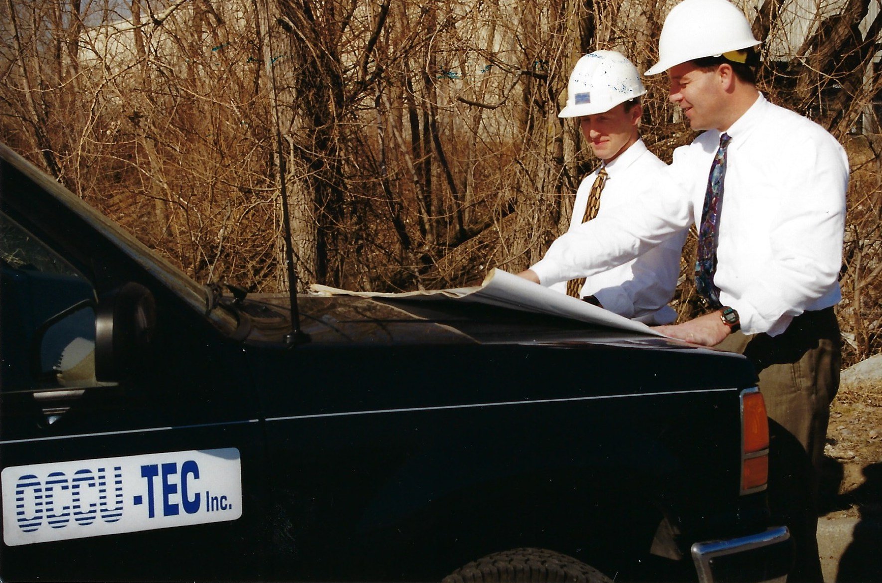 Two men in white shirts and ties with white hard hats inspect paper plans on the back of a 1980s car.